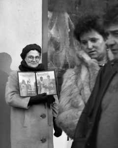 An elderly lady stands holding an open copy of The Watchtower, a magazine published by The Jehovah's Witnesses, a Christian sect, on the pavement in Augsburg, Germany, as a heterosexual couple passes by.